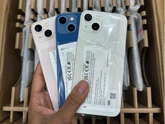 iPhone 13 lacrados nas cores rosa, azul e branco sobre uma caixa de atacado